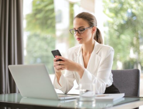 Concentrated young woman in formal clothes and eyeglasses sitting at glass table with laptop while surfing smartphone