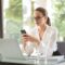 Concentrated young woman in formal clothes and eyeglasses sitting at glass table with laptop while surfing smartphone