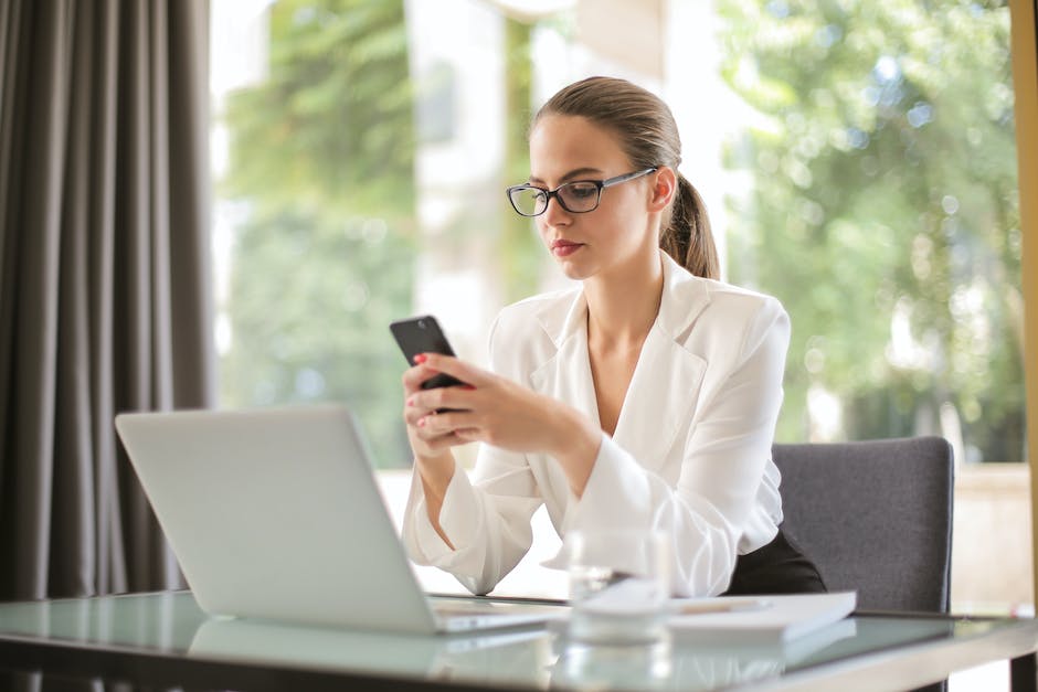 Concentrated young woman in formal clothes and eyeglasses sitting at glass table with laptop while surfing smartphone
