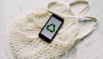 Overhead of smartphone with simple recycling sign on screen placed on white eco friendly mesh bag on marble table in room