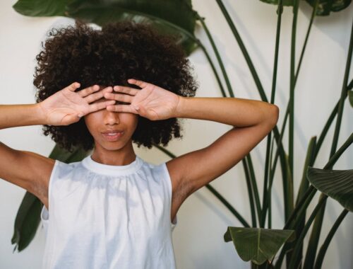 African American female with Afro hairstyle standing near green plant and hiding eyes behind hands