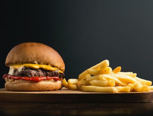 Appetizing burger with meat patty ketchup and cheese placed on wooden table with crispy french fries against black background
