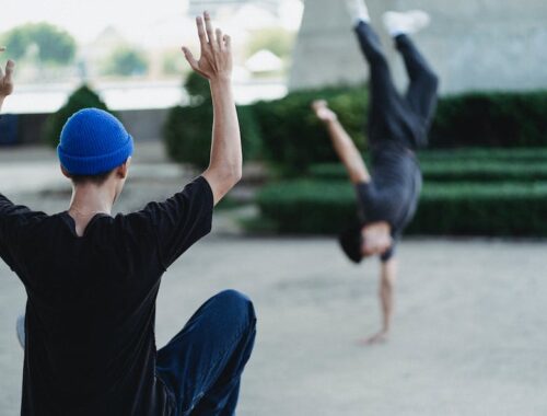 Back view of faceless male in casual clothes instructing friend while performing handstand on city street