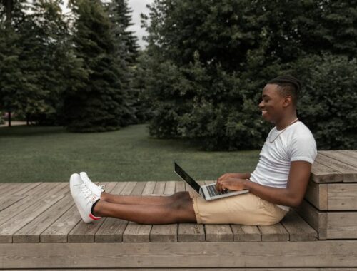 Side view of African American male in casual outfit sitting on wooden surface and browsing netbook while looking at screen