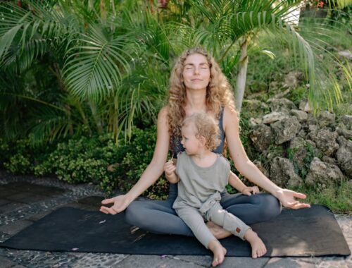 Full body happy relaxed mother sitting with eyes closed in lotus pose with cute boy on laps while practicing yoga in tropical green park