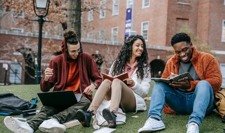 A diverse group of happy students sitting on a grassy university lawn, using notebooks and a laptop while studying together.