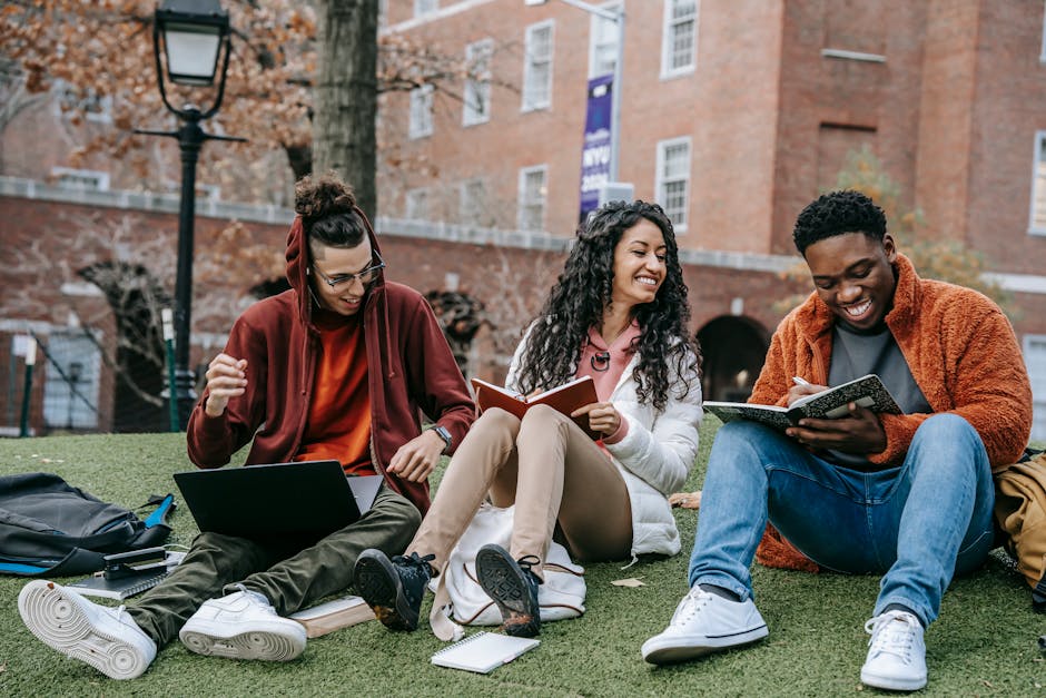 A diverse group of happy students sitting on a grassy university lawn, using notebooks and a laptop while studying together.